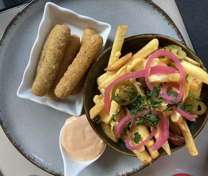 En tallrik med mozzarellasticks i en skål, cheezy fries med jalapeños och picklad rödlök, toppad med örter, samt en klick dipp på sidan.