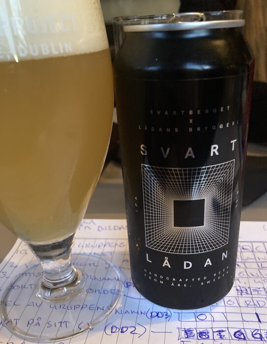 Ett glas öl och en burk med etiketten "Svart Lådan" från Svartberget x Lådans Bryggeri, står på ett papper med skrivna anteckningar.