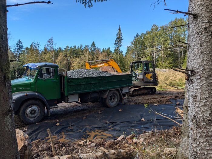 En grön lastbil med släp fylld med stenar för parkering, en grävmaskin bredvid, omgiven av skog.