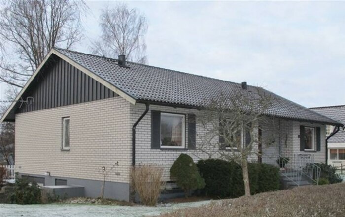 Ett enplanshus från 60-talet med mexitegelfasad och stående träpanel på gaveln. Huset har nya stora fönsterpartier och vegetation runtomkring.