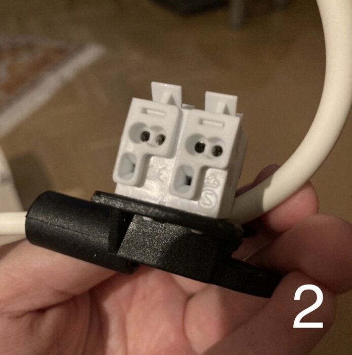 Närbild på en hand som håller en vit sockerbit för elinstallation med en vit kabel ansluten, markant nummer två synligt.