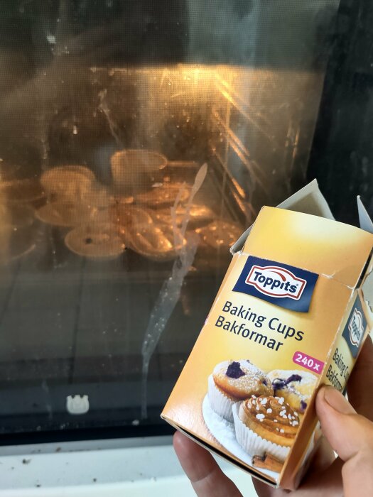 Förpackning med bakformar från Toppits hålls framför en ugn med överflödande muffinsformar.