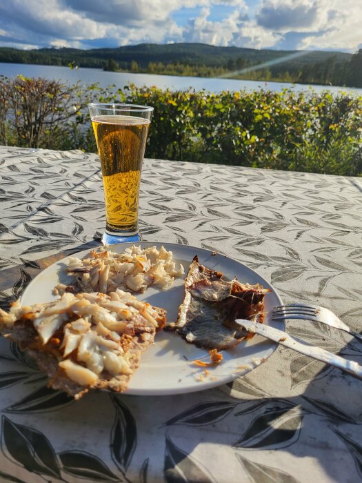 Tallrik med två knäckebrödskivor toppade med smörstekta kantareller och rökt abborre, tillsammans med ett glas öl, utomhus vid en sjö med utsikt över landskapet.