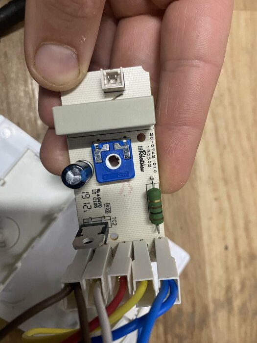 Närbild av en hand som håller en vit elektronisk termostatkomponent med diverse färgade kablar anslutna till en frysbox.