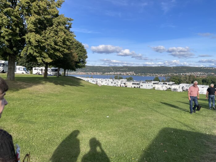 Grön park med husbilar parkerade nära vatten, några personer promenerar i förgrunden under en klarblå himmel med vita moln.