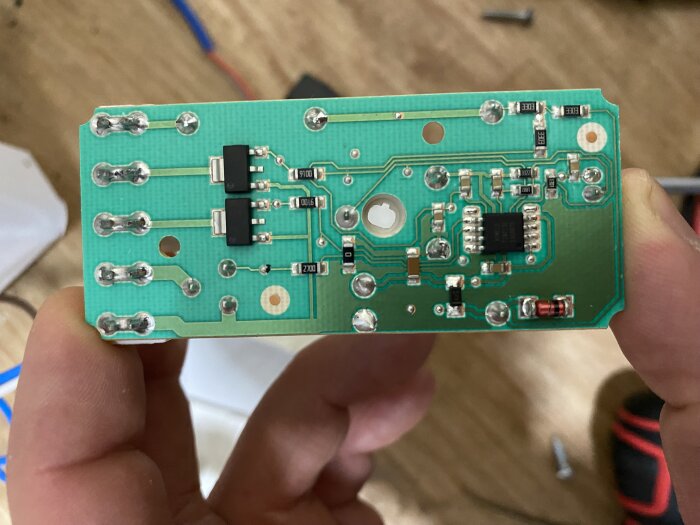 Närbild på ett grönt kretskort med olika elektroniska komponenter, hål och kopparledningar. Kretskortet hålls upp för att visa dess detaljer.