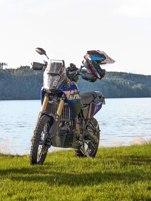 Blå motorcykel på gräs vid en sjö med skog i bakgrunden, hjälm placerad på styret, fotot är taget under klarblå himmel.