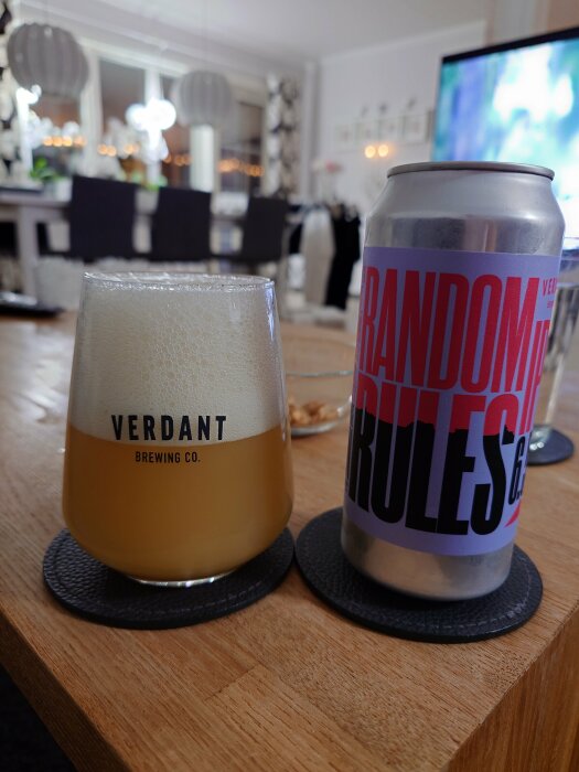 En öl från Verdant Brewing Co. i ett glas bredvid en burk med etiketten "Random Rules", står på ett träbord i ett modernt inrett rum.