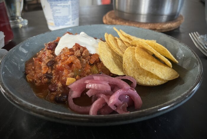 En tallrik med chili con carne med grädde, picklad rödlök och nachos.