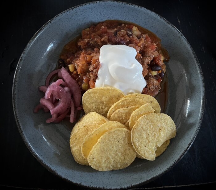 En tallrik chili con carne med grönsaker, toppad med gräddfil, picklad rödlök och nachos vid sidan.