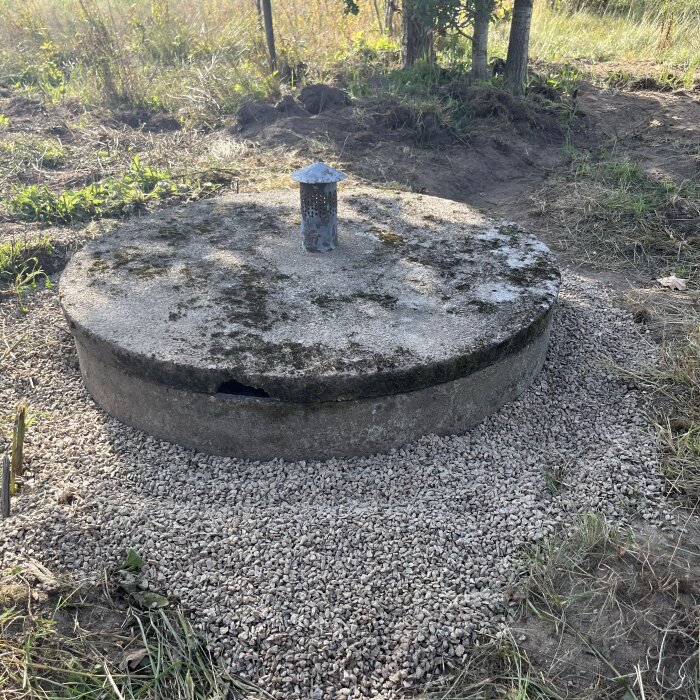 En grävd brunn med betonglock, synligt slitage och skada, två infasningar för grepp. Området runtomkring är fritt från vegetation och täckt med grus.