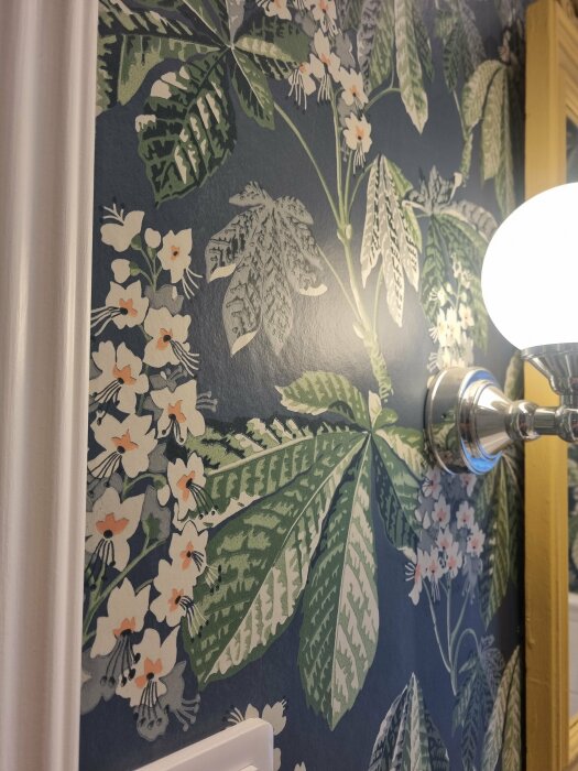 Close-up of a dark blue wallpaper with green leaves and white flowers, illuminated by a lamp on the right side.