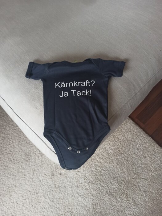 Mörkblå babybody med texten "Kärnkraft? Ja Tack!" tryckt på ryggen, liggande på en ljus soffa.