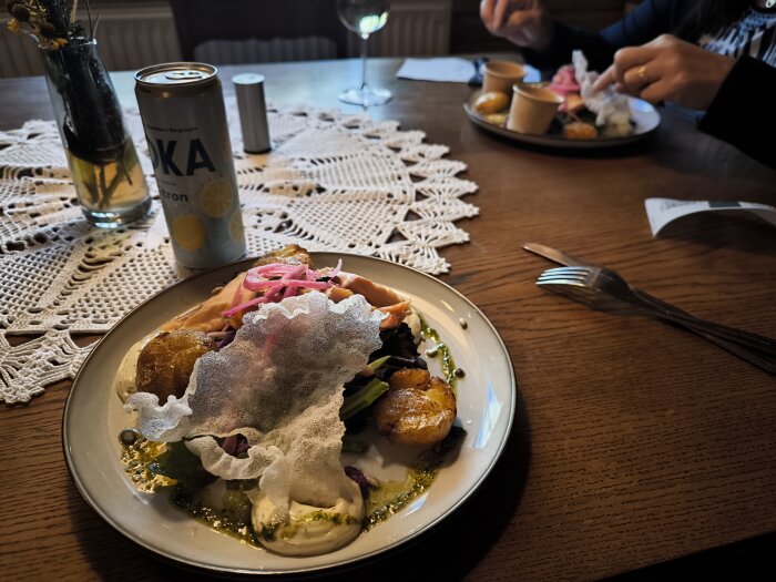 Tallrik med rökt lax och diverse tillbehör, en burk dryck och en annan tallrik i bakgrunden på ett träbord med vit duk.