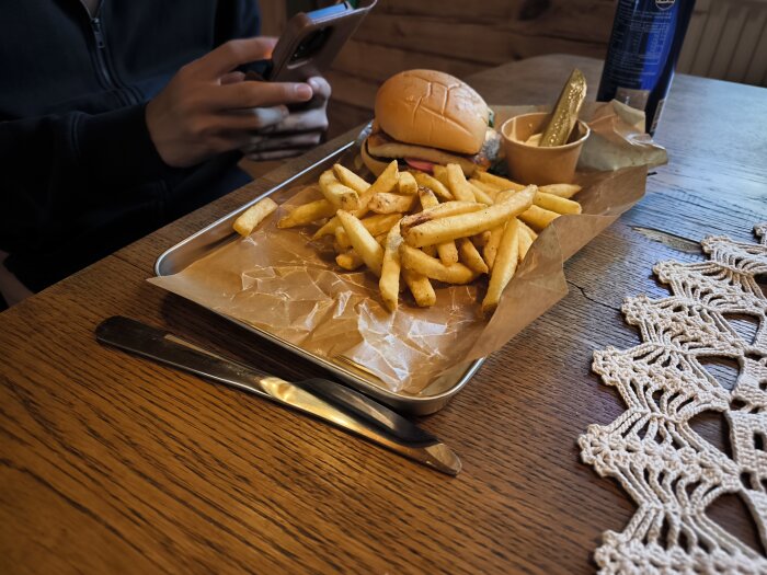 En hamburgare med halloumi och pommes frites ligger på en bricka med brunt papper, bredvid syns en burk med dryck. En person håller en mobiltelefon.