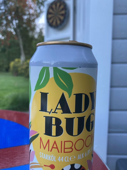 En burk med öl av märket "Ladybug Maibock" står på ett bord utomhus med en darttavla i bakgrunden.