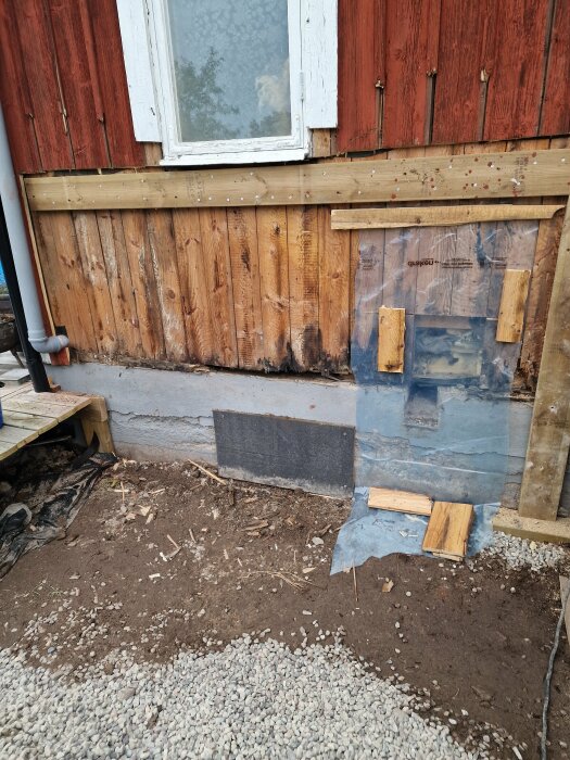 Pågående renovering av fasad med skadad panel och syll under ett fönster, byggmaterial och grus framför huset.