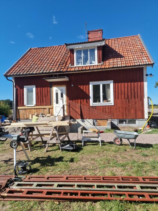 Rött hus med röda takpannor, byggställningar och verktyg utanför. Pågående renovering med byggmaterial och verktyg runt omkring. Klarblå himmel.