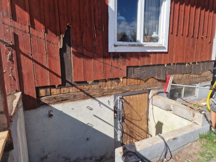 Halvfärdigt fasadrenoveringsprojekt på ett rött hus, där en del av träpanelen är borttagen och väggen blottlagd. En kvast lutandes mot väggen vid fönstret.