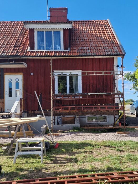 Ett hus under renovering med byggställningar framför fasaden, där träpaneler delvis har tagits bort och byggmaterial ligger utanför huset.