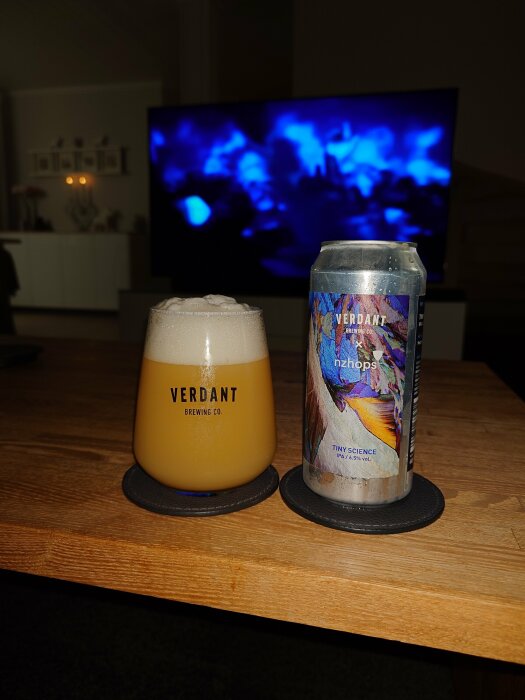 Ölglas och ölburk från Verdant Brewing Co. på ett bord, med en TV som spelar en blåfärgad scen i bakgrunden.