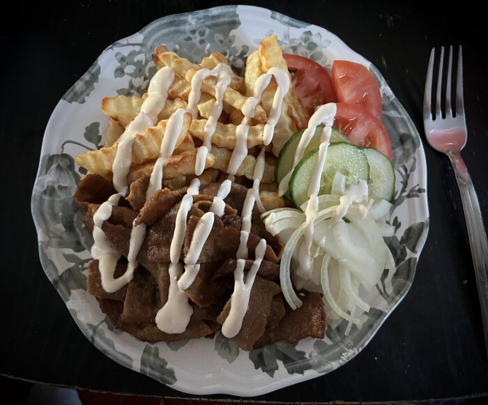 Tallrik med stekt kebabkött, pommes frites, skivad tomat, gurka och rå lök, toppad med vitlöksdressing.