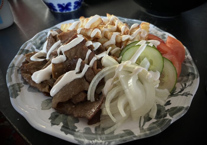 Tallrik med kebabkött, pommes frites, tomat, gurka, rå lök och vitlöksdressing.