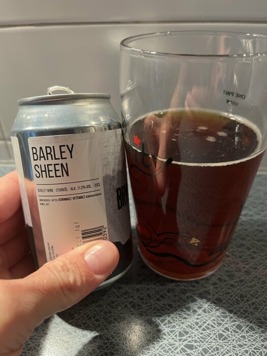 Hand håller en burk "Barley Sheen" öl bredvid ett glas fyllt med mörk öl. Text på burken beskriver ingredienser och alkoholhalt.