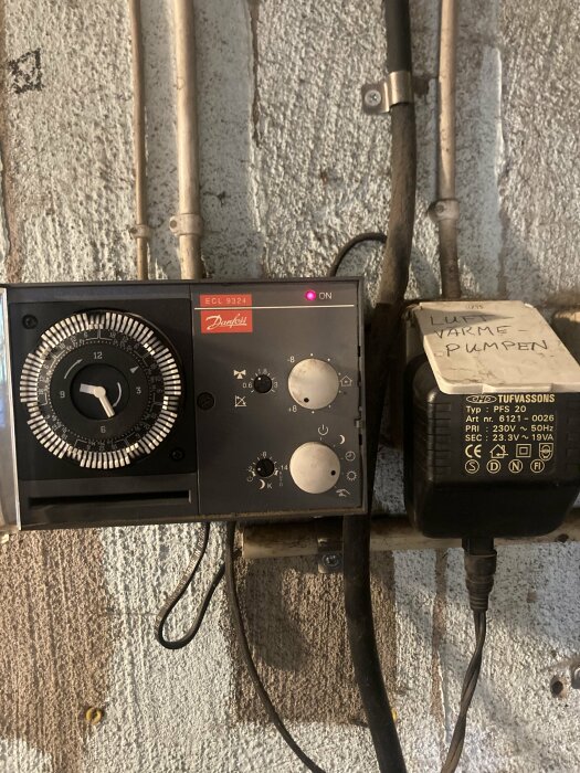 Danfoss ECL 9324 regulator monterad på väggen med inställningsvred och lampa som lyser. En märkning "Lur VARME PUMPEN" syns på en kopplingsbox intill.