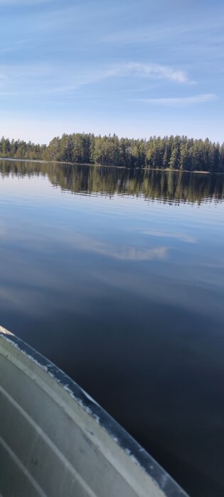 Visar en stilla sjö med spegelblankt vatten, omgiven av en skogsstäckt strandlinje under klarblå himmel; kanten av en båt syns i förgrunden.