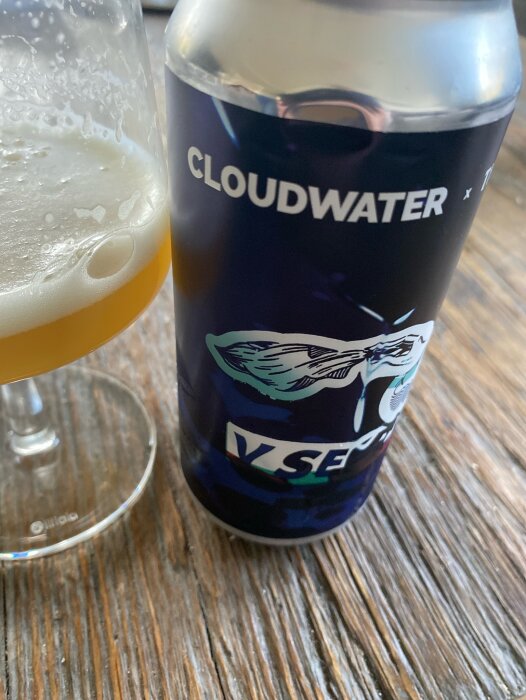 En burk Cloudwater The Veil Brewing co öl bredvid ett halvfullt glas med skummig öl på ett träbord.