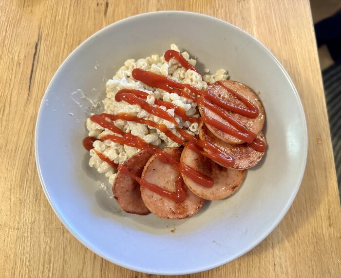 En tallrik med makaroner och stekt falukorv toppat med ketchup på ett bord av trämaterial.