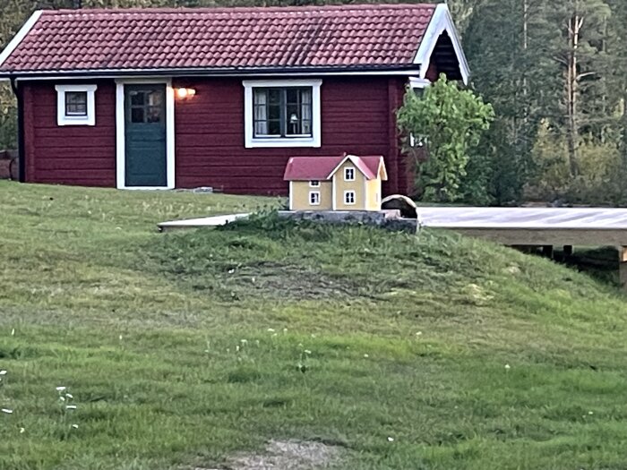 Rött hus med vita knutar och gröna dörrar i bakgrunden, och en miniatyr av ett gult hus med rött tak framför, omgivna av gräs och träd.