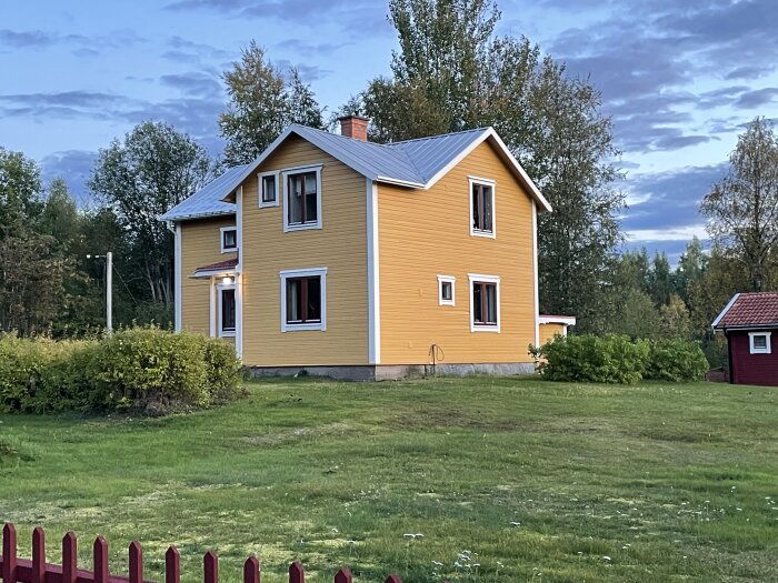 Gult tvåvåningshus med vitmålade fönsterkarmar på en grön gräsmatta, omgiven av träd och buskar, under molnig himmel på kvällen.