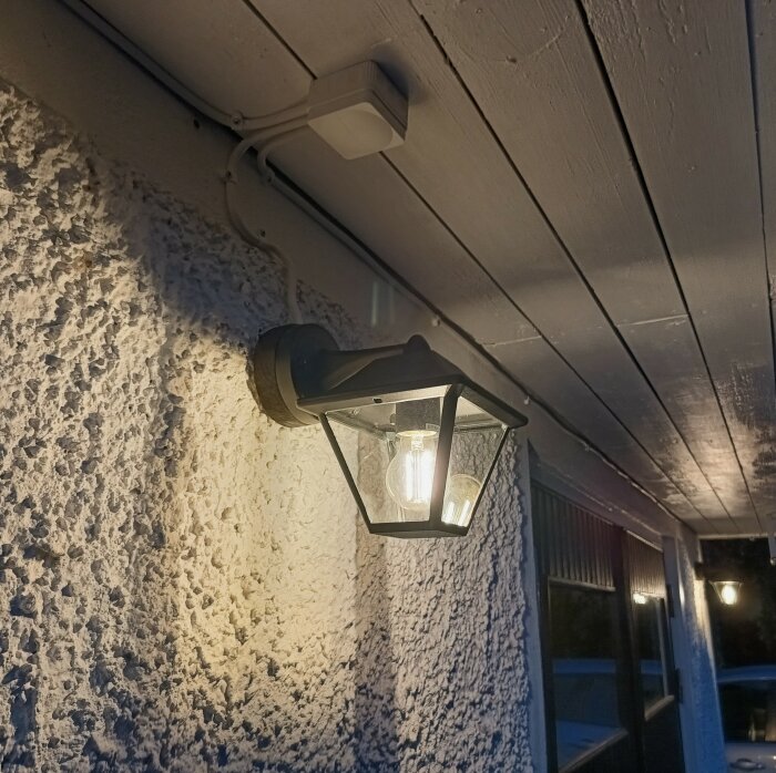 Nyinstallerad svart vägglampa på garagefasad under takfoten, med lysande glödlampa inuti.