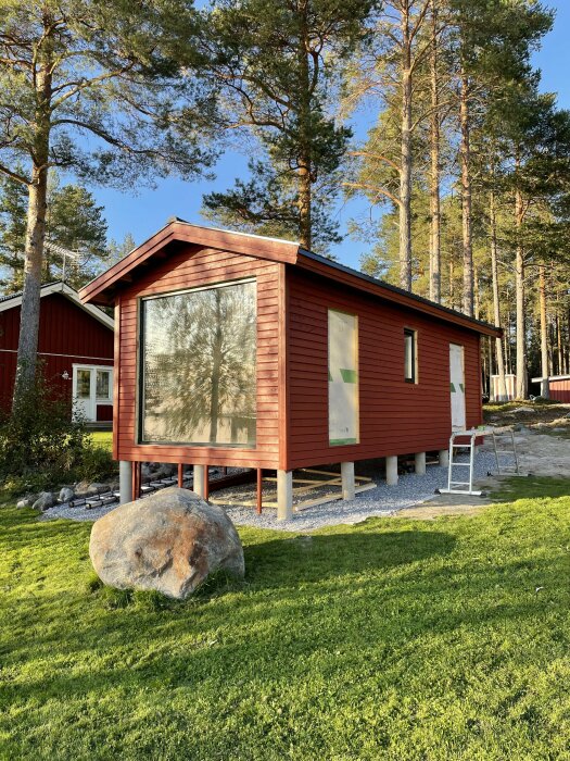 Ett rött fritidshus med stora fönster är nyrenoverat och omgivet av skog. En stege står lutad mot väggen och en stor sten finns på gräsmattan framför.