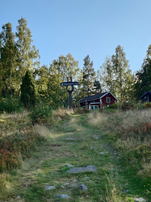 En gångstig leder upp till en röd stuga bakom en skylt som säger "Fäbodvallen" med träd i bakgrunden.