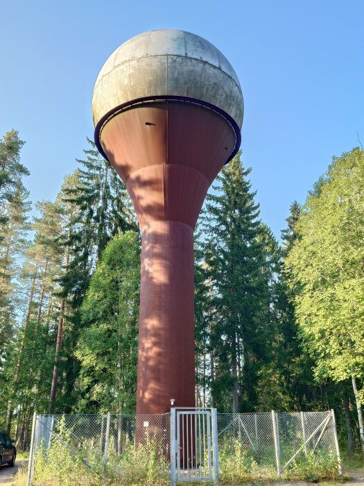 Vattentorn med rund betongbehållare ovanpå en hög pelare, omgiven av stängsel och skog i bakgrunden.