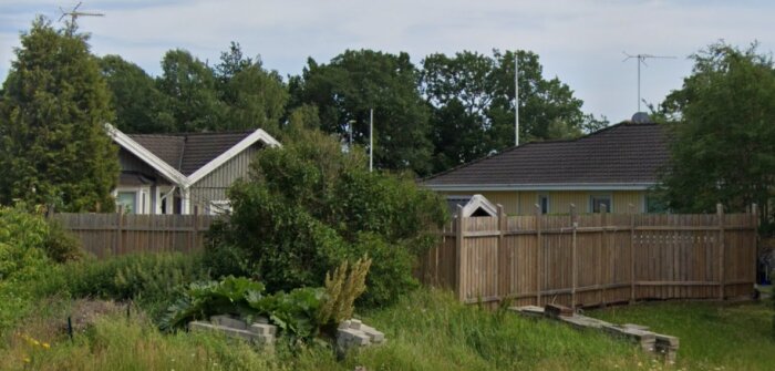 Två hus med en hög plankstängsel mellan sig, ett med förråd eller poolhus, omgivna av trädgård och grönska i ett bostadsområde.