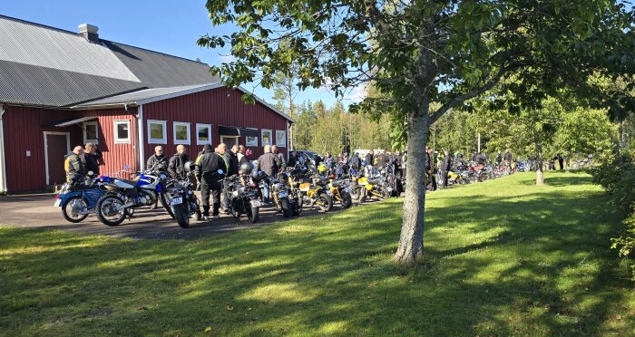 En grupp deltagare i ett mopedrally samlade utanför en röd byggnad med sina mopeder i det soliga vädret. Träden och gräset omger området.