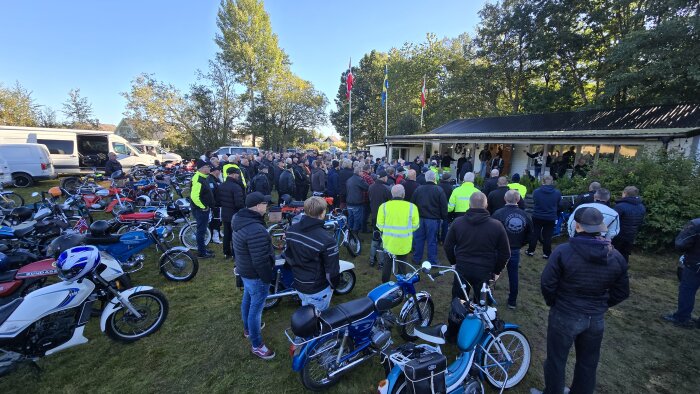 En stor samling människor, några i reflektionsvästar, står utanför en byggnad omgiven av mopeder. Det är en solig dag med träd i bakgrunden och flaggor som vajar.