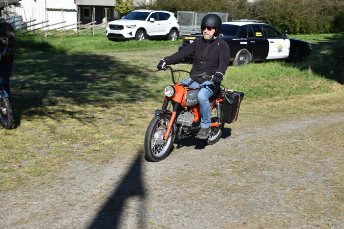 Person i svart hjälm kör en orange Zundapp-moped på en grusväg under mopedrally med gräs och parkerade bilar i bakgrunden.