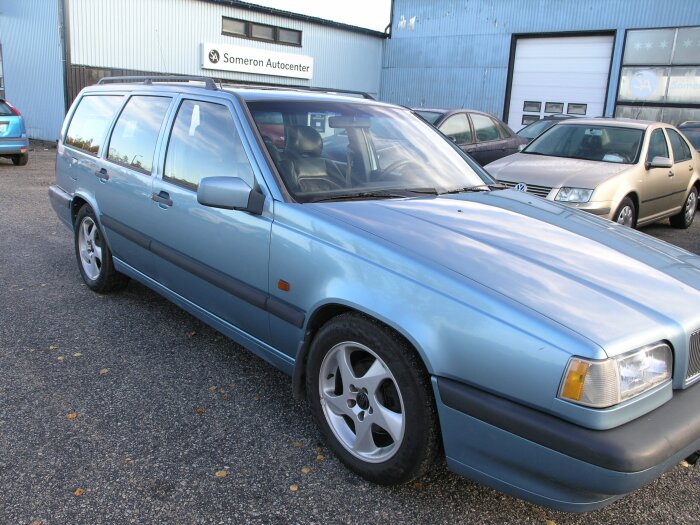 Blå Volvo 850 kombi parkerad framför en bilverkstad med andra bilar i bakgrunden.