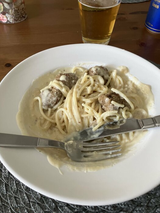 En tallrik pasta med gorgonzolasås och köttbullar, en gaffel och en kniv, samt ett glas öl på ett träbord.