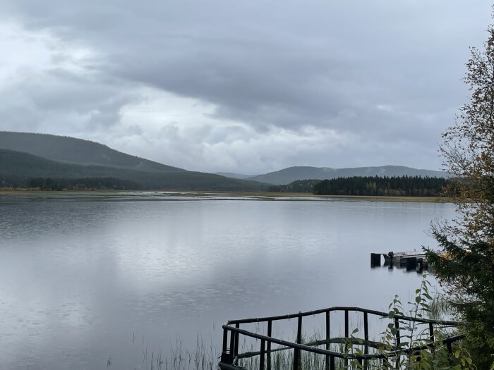 En lugn sjö med en liten brygga, omgiven av skogbeklädda kullar under en molnig himmel.