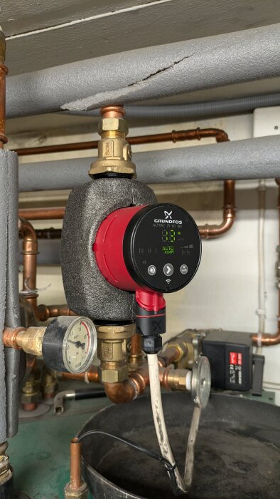 Nyinstallerad Grundfos cirkulationspump av modell ALPHA2 25-60 180 ansluten till rörsystem, med en tryckmätare och diverse rör och kopplingar i bakgrunden.