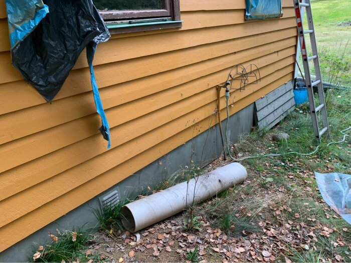 Gul trästuga med synlig krypgrund, en trädgårdsslang och en mindre stege står lutad mot väggen. Plastskynken täcker delvis fönstren och viss vegetation syns på marken.