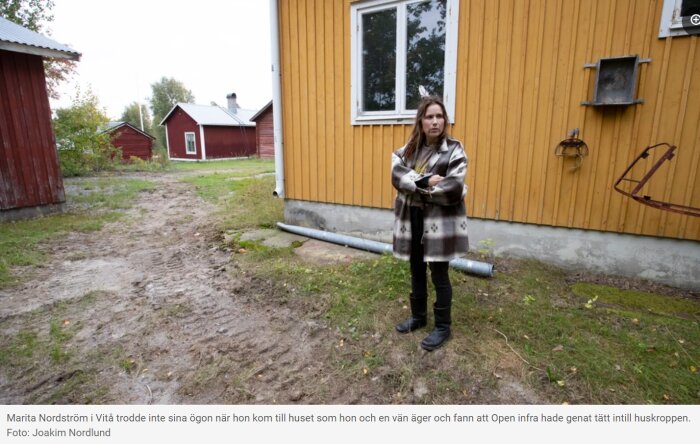 Kvinna står med armarna korsade bredvid ett gult hus, omgiven av flera röda byggnader och en uppgrävd jordig mark med hjulspår.