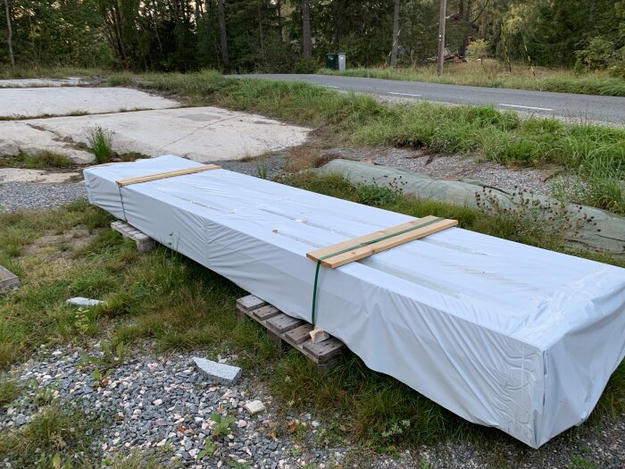 Plastinpackad innerpanel ligger på träpallar utomhus vid en grusgång med gräsmatta i bakgrunden och en landsväg.