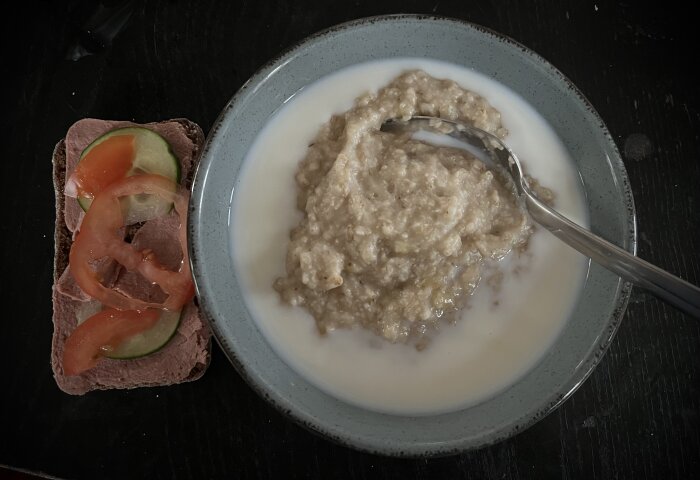 En skål med havregrynsgröt i mjölk bredvid en leverpastejsmörgås med gurka och tomat.